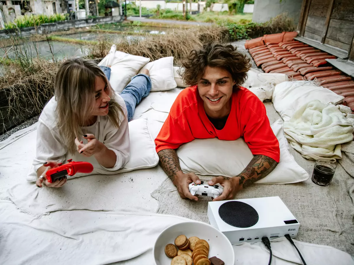 A couple is laying down on a patio, playing video games on a white PS5 console. The man is in a red shirt and the woman is in a white shirt.  They are smiling and looking at each other. There are snacks and drinks on the patio.