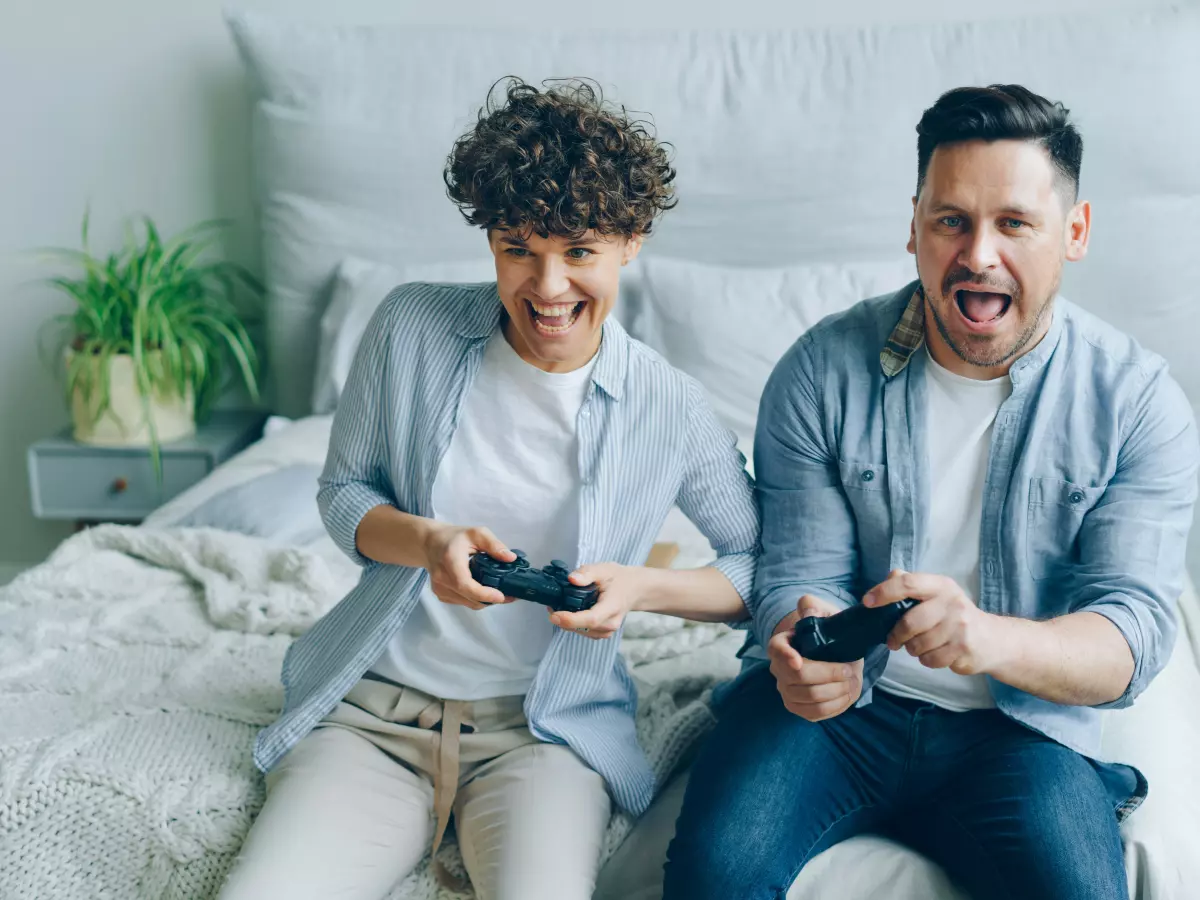 A couple playing video games on a bed.  The man and woman are both looking at the camera with their mouths open in surprise.  The woman is holding the controller.  They are both wearing casual clothing.