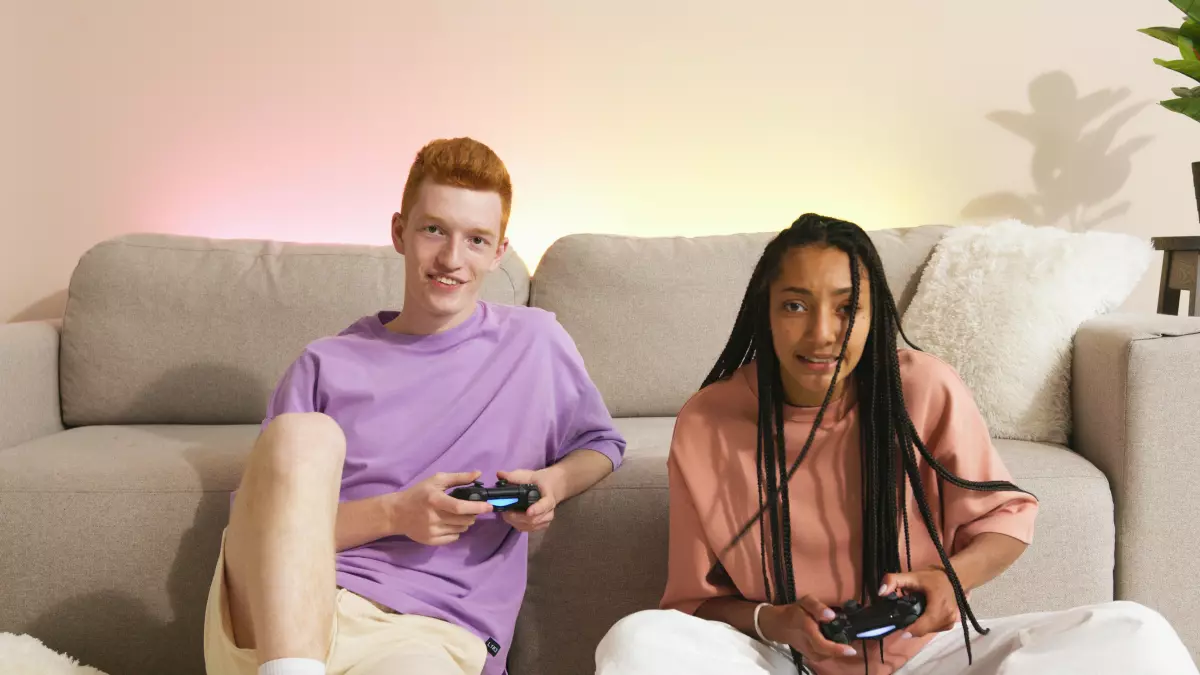 Two young adults, a man and a woman, are sitting on a couch, each holding a game controller. They are smiling and appear to be having fun playing a video game together.