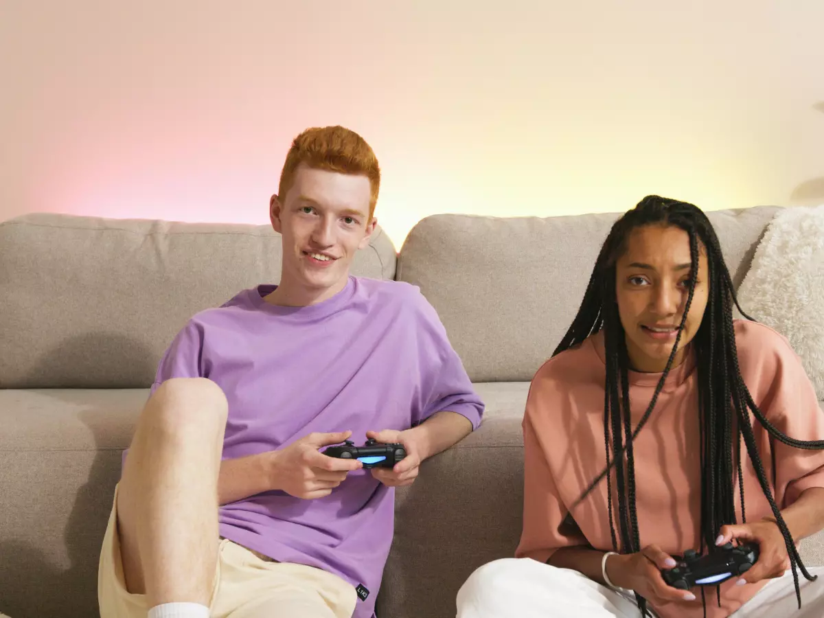 Two young adults, a man and a woman, are sitting on a couch, each holding a game controller. They are smiling and appear to be having fun playing a video game together.