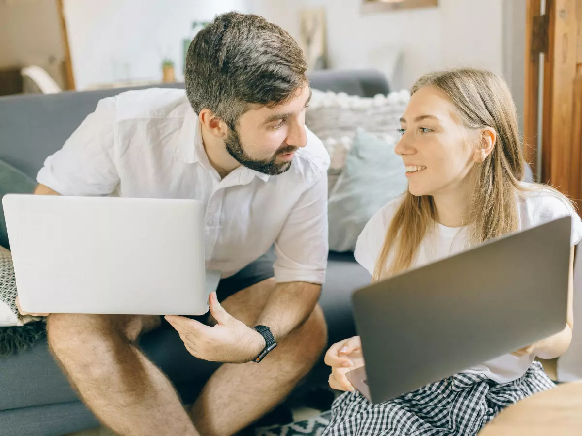 A man and a woman are sitting on a couch, each with a laptop in their laps. They are looking at each other.