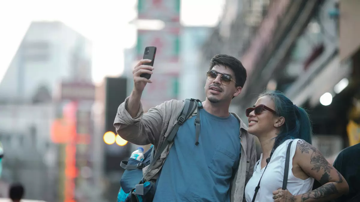 A person takes a selfie with a phone in the street with another person in the background.