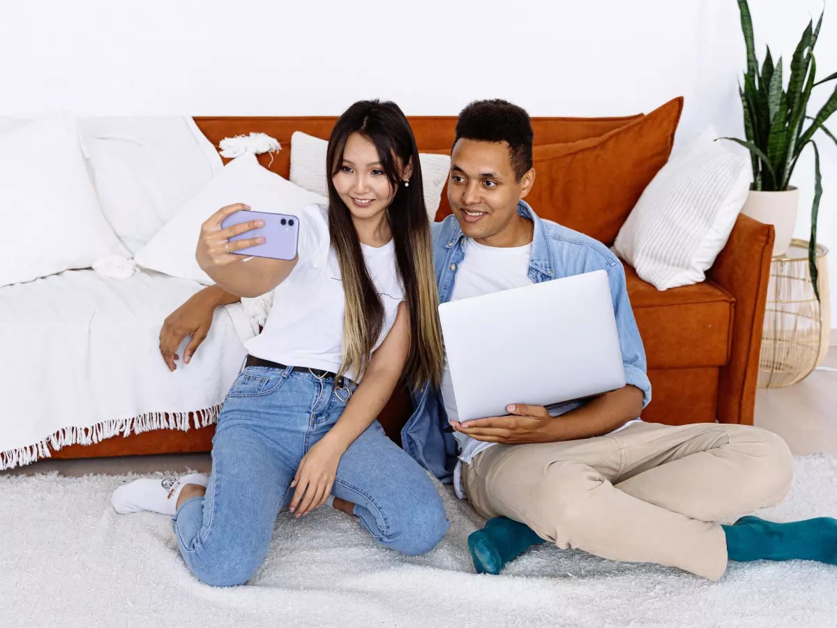 A woman and a man are sitting on the floor, looking at a laptop screen. The woman is holding a smartphone and smiling. The man is looking at the laptop screen with interest.