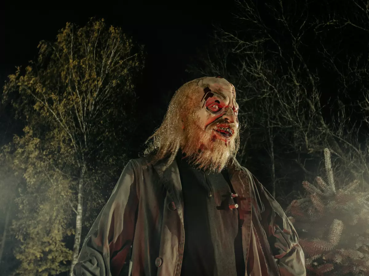 A zombie with a grotesque mask and long hair is standing in a forest setting. The zombie is looking at the camera with a menacing expression.