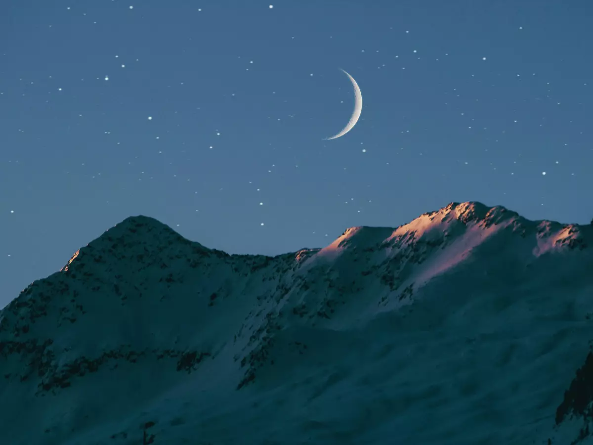 A crescent moon hangs in the night sky above a mountain range.