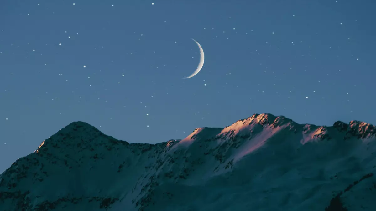 A crescent moon hangs in the night sky above a mountain range.