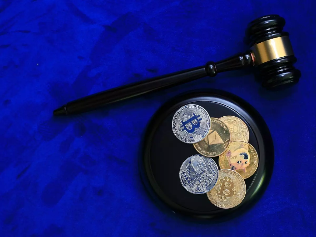 A judge's gavel lies on a black surface with a group of cryptocurrency coins on a blue background.