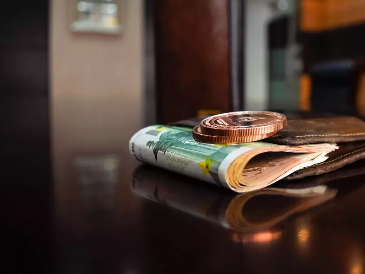 A brown leather wallet with euro bills and a pair of crypto coins on a reflective surface.