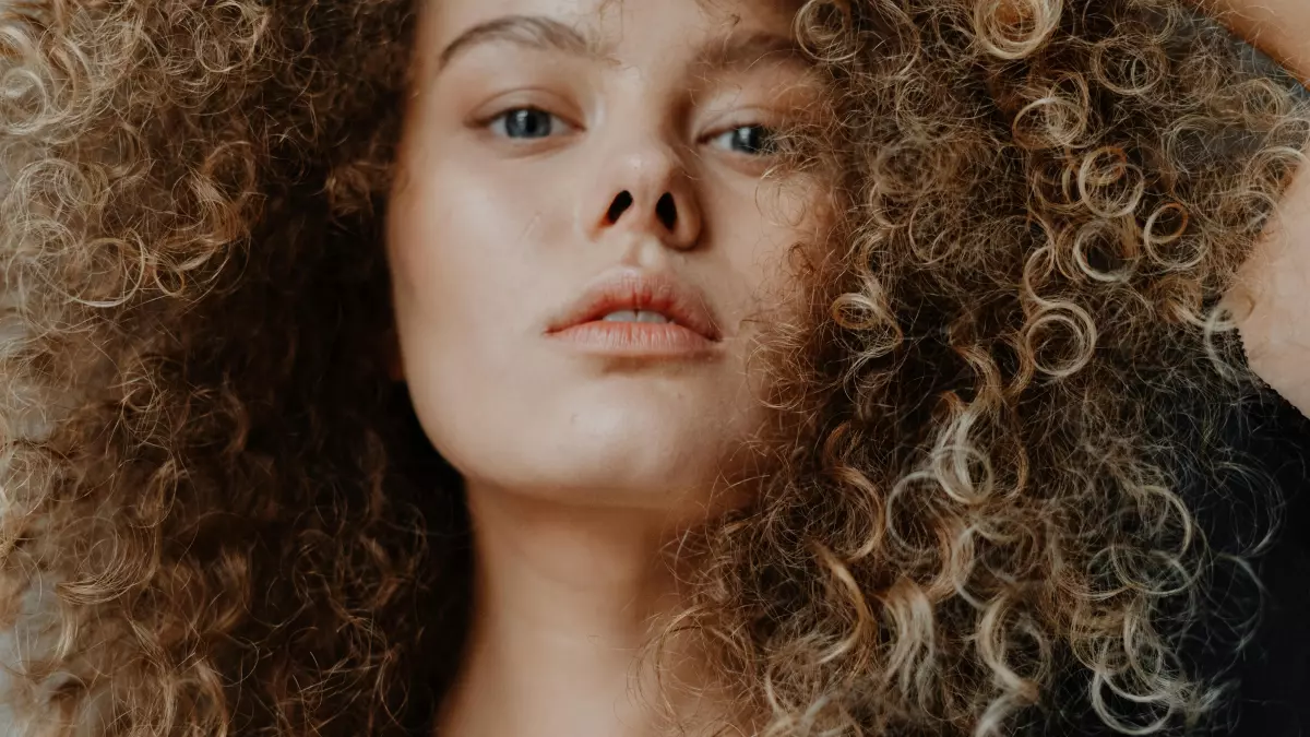 Close-up of a young woman with thick, curly, light brown hair. Her expression is serious.