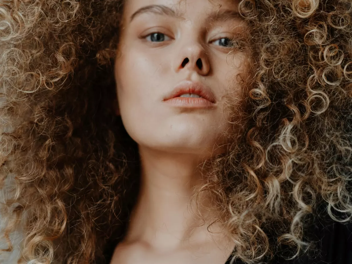 Close-up of a young woman with thick, curly, light brown hair. Her expression is serious.