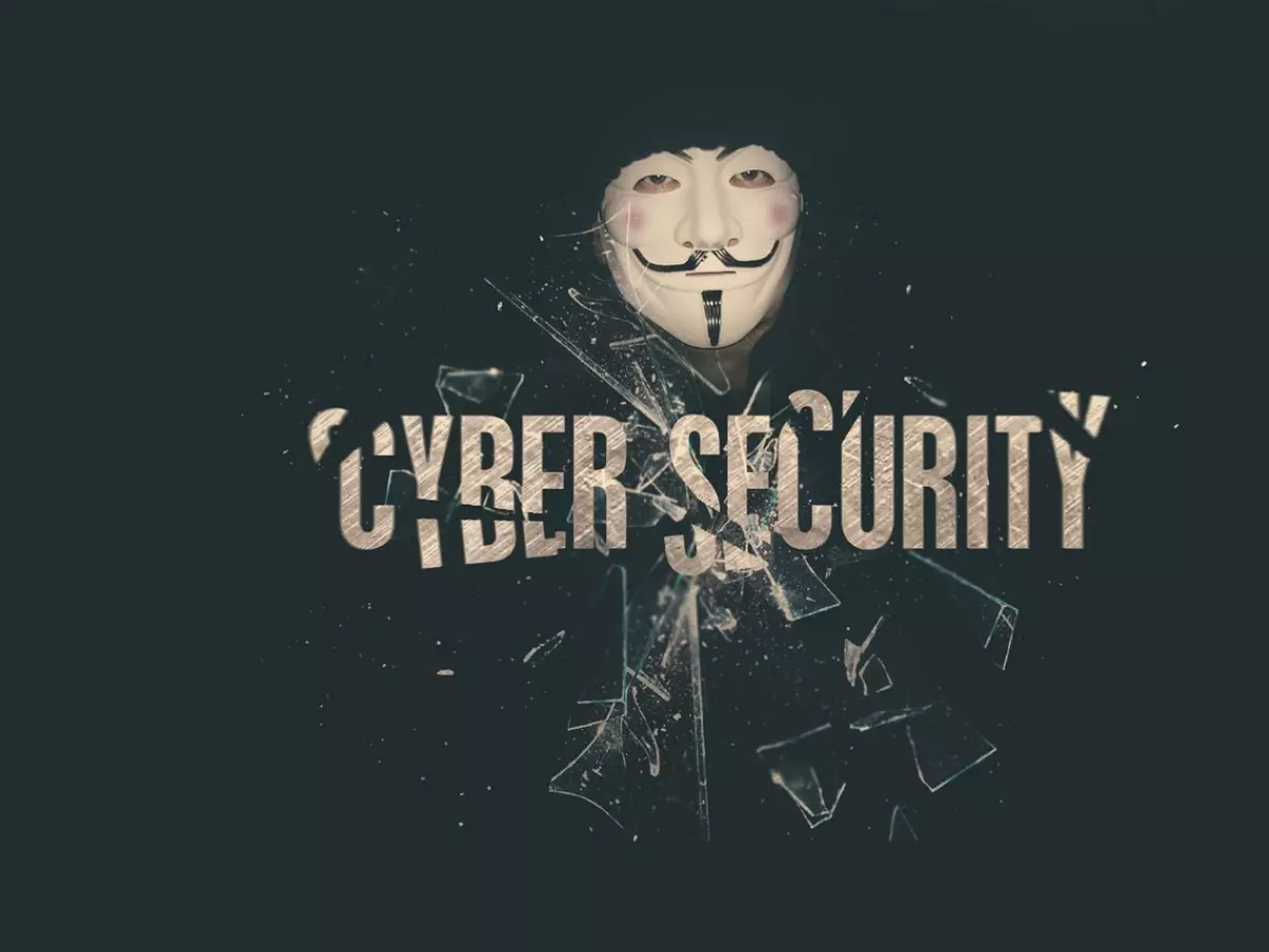 The image is a dramatic portrayal of a man wearing a mask. The mask is the famous Guy Fawkes mask. He is partially concealed by broken glass shards. The word cybersecurity is placed on top of broken glass shards. The background is dark, which adds to the mysterious and ominous tone.