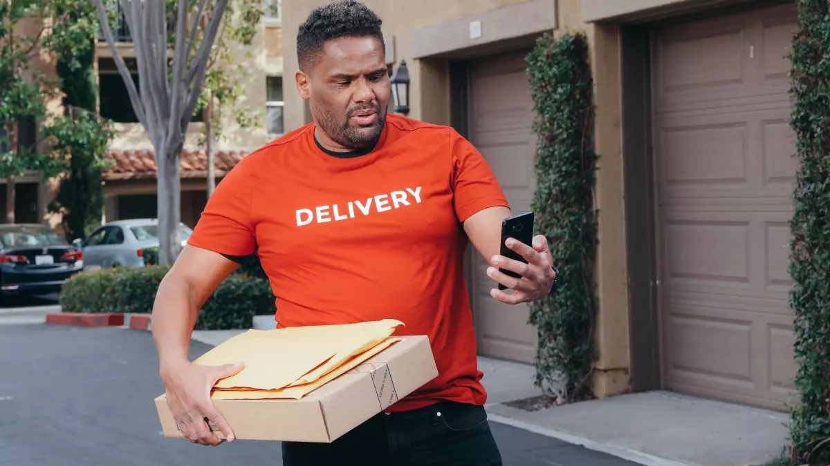 A man in a red delivery uniform, holding a package in his arms, is looking at his smartphone in shock, with a look of surprise on his face. He is standing in front of a house in a residential area, with a garage door in the background.