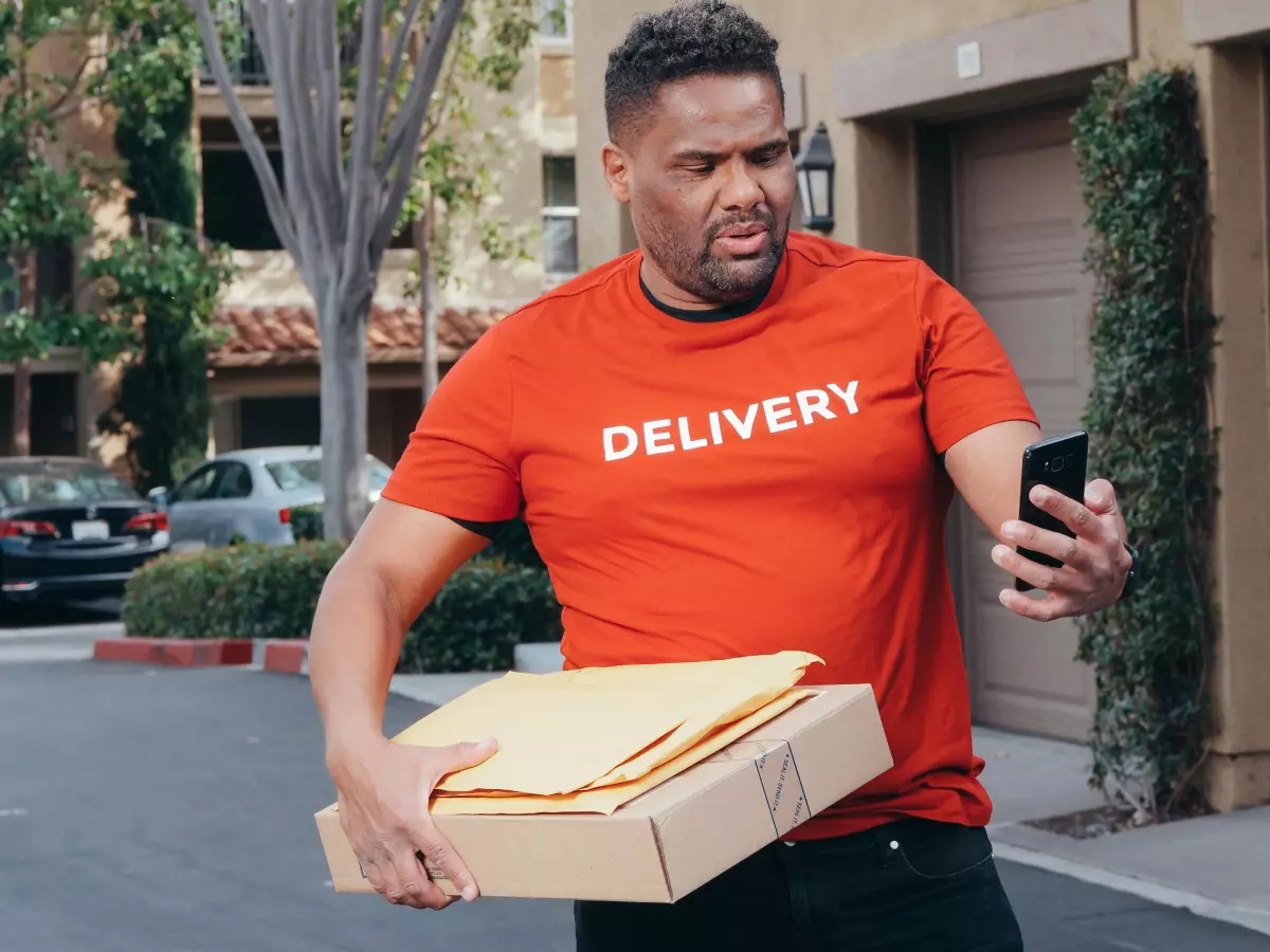 A man in a red delivery uniform, holding a package in his arms, is looking at his smartphone in shock, with a look of surprise on his face. He is standing in front of a house in a residential area, with a garage door in the background.