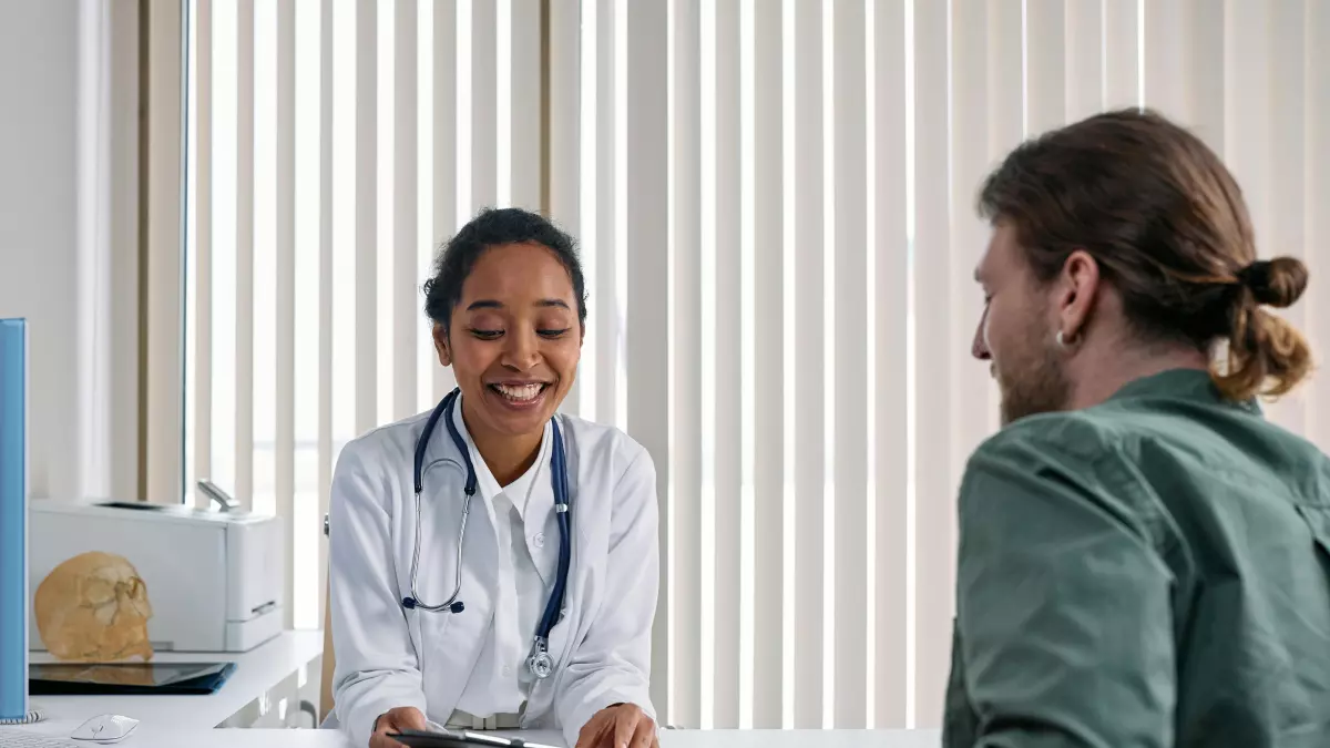 A female doctor in a white coat is sitting at a desk, smiling and looking at a male patient. She is holding a clipboard in her hand.