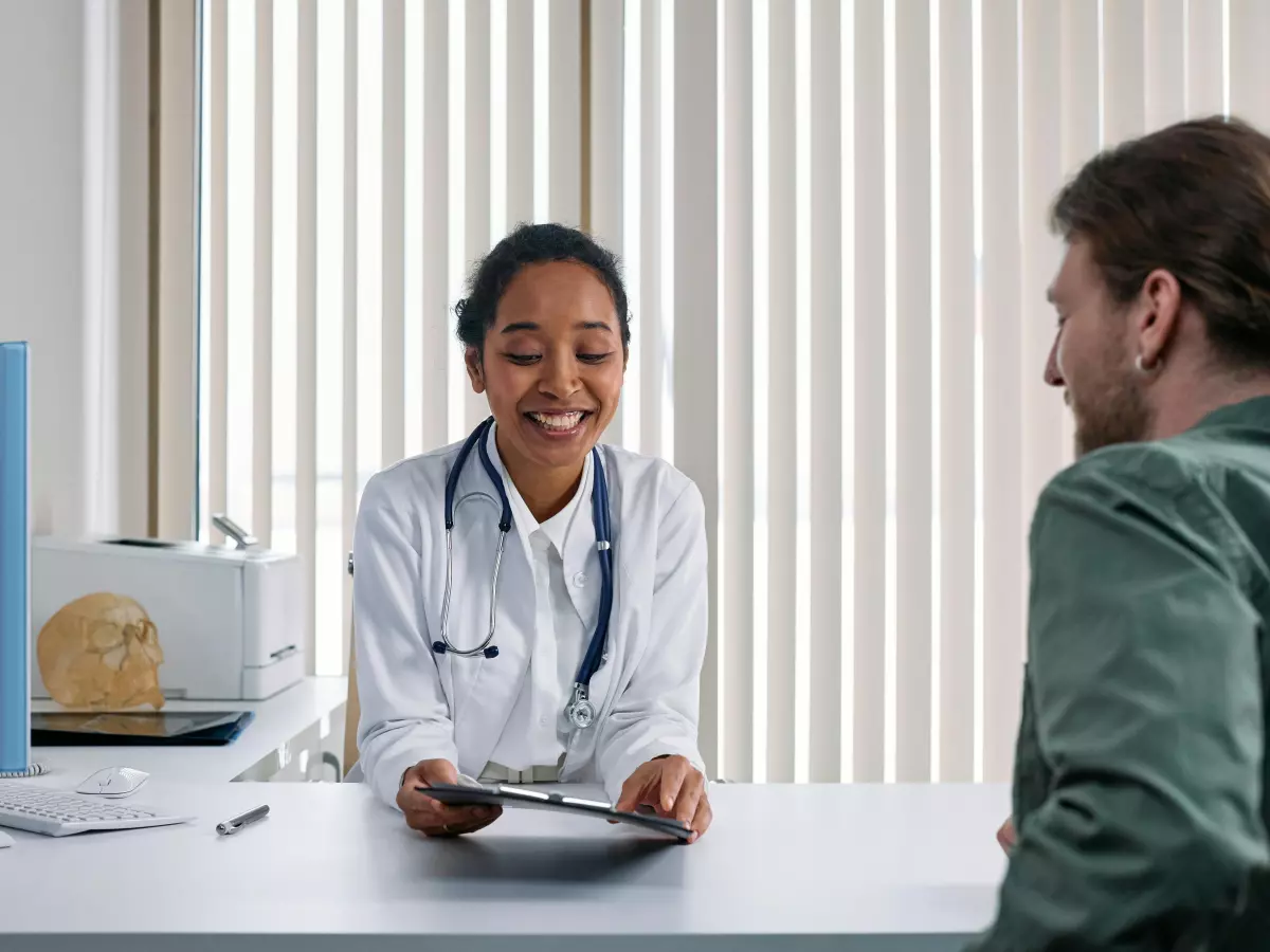 A female doctor in a white coat is sitting at a desk, smiling and looking at a male patient. She is holding a clipboard in her hand.