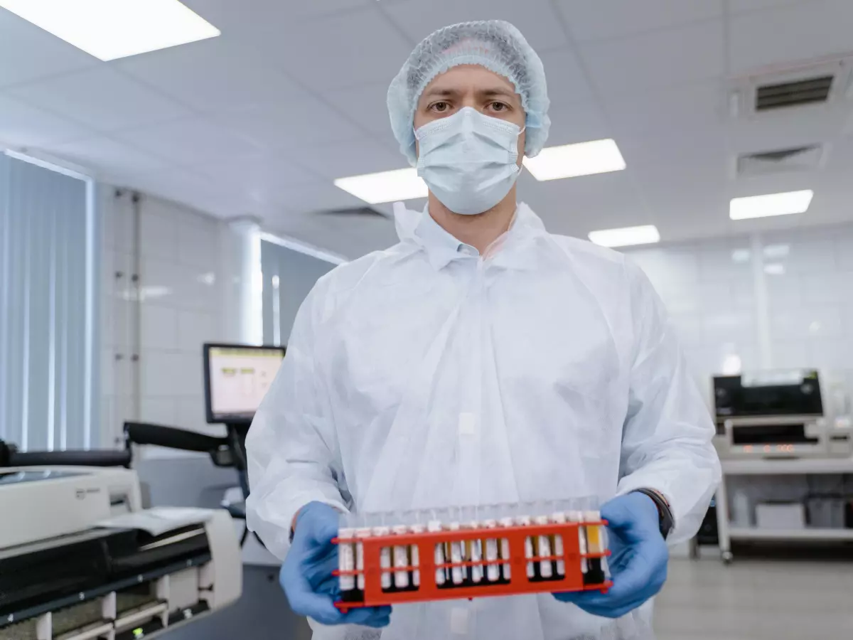 A scientist in a lab setting holds a tray of test tubes, looking directly at the camera.