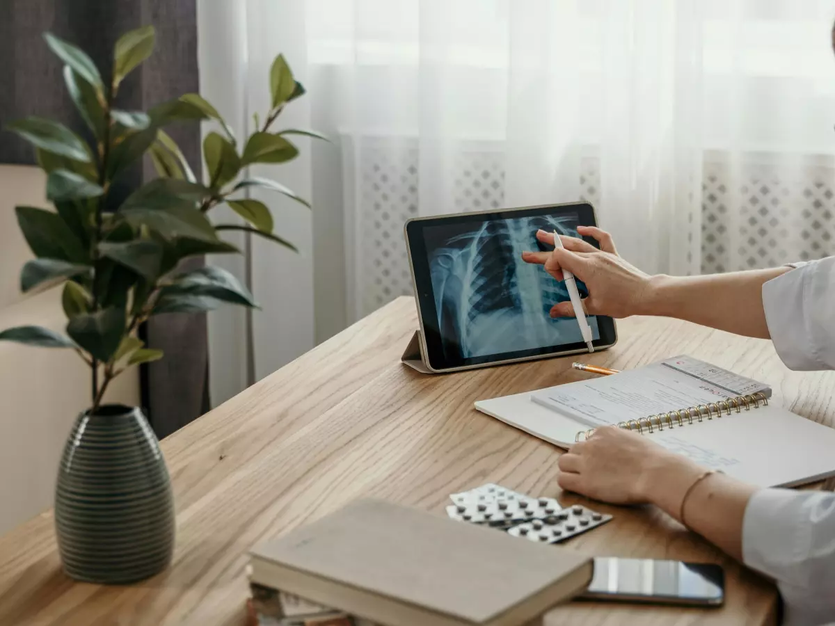 A woman sitting at a desk looking at a tablet computer while holding a pen. There is a plant and papers on the desk and an X-ray image on the screen.