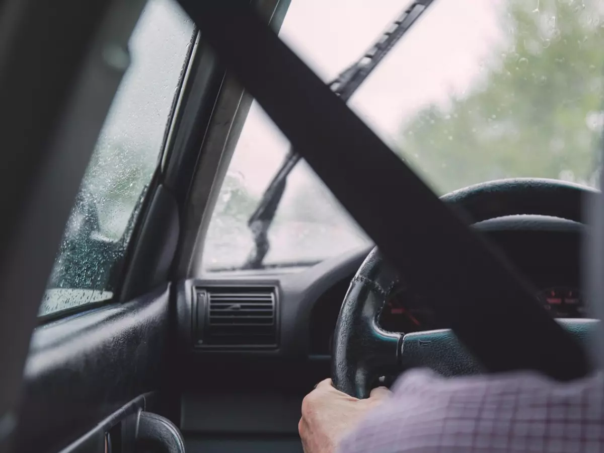 A driver's hand on the steering wheel, with a seatbelt visible, driving through a rainy street.