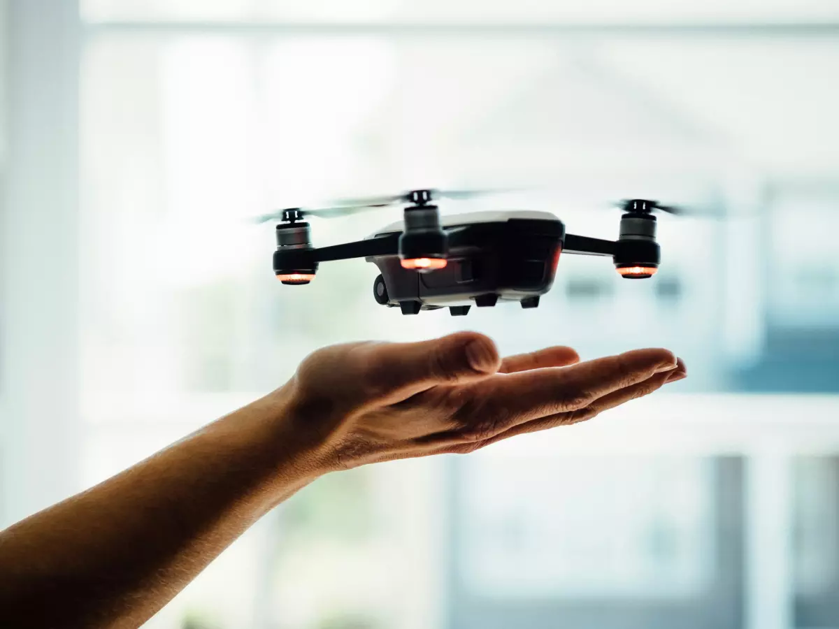 A close-up photo of a drone hovering above a person's outstretched hand. The drone has four propellers and a small camera attached to the bottom.