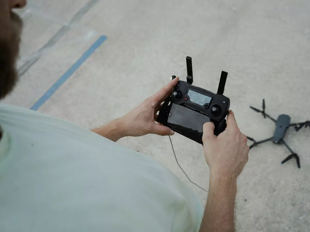 A man's hands holding a drone remote control. The drone is in the background, and it is partially visible.