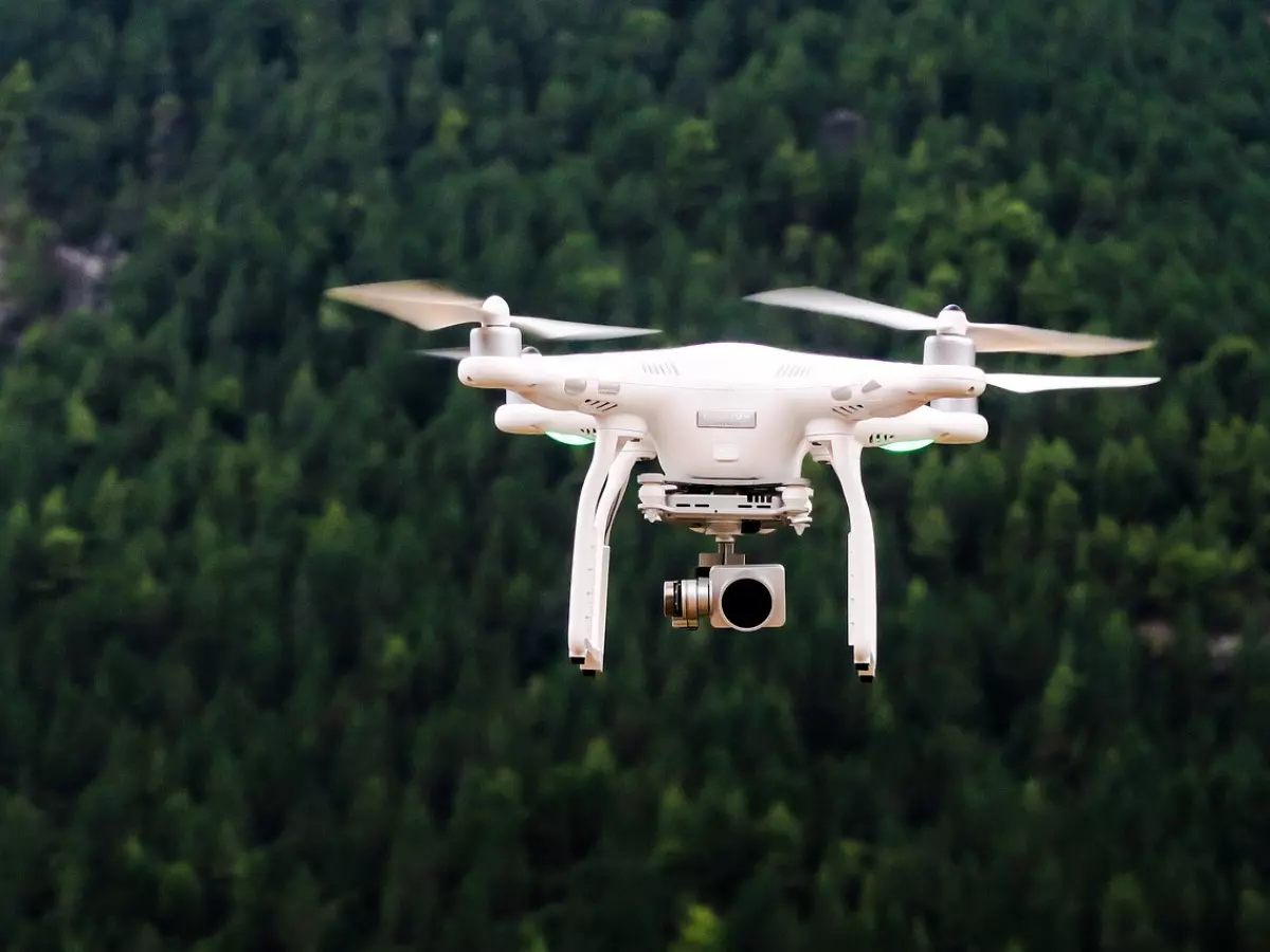 A white drone flying over a green forest. It's a clear sunny day and the drone is in mid-flight.