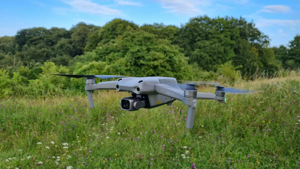 A drone is in flight, viewed from the front, with the camera lens visible. It is in front of a blurry background of trees.