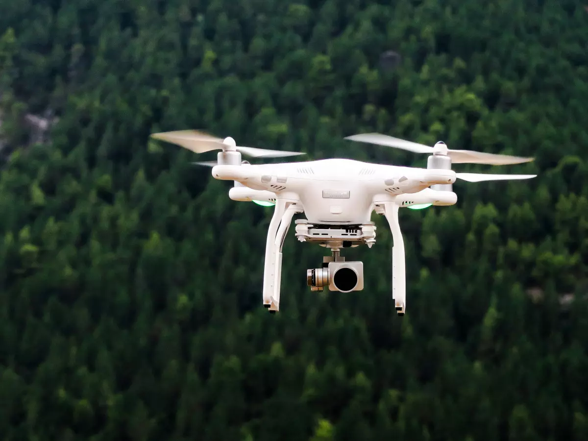 A white drone is flying in the air with a green forest in the background.