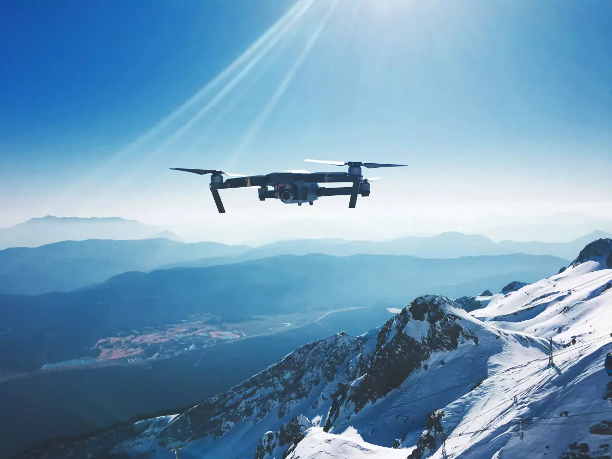 A drone flies high above a snowy mountain range, with a clear blue sky and sunbeams in the background.