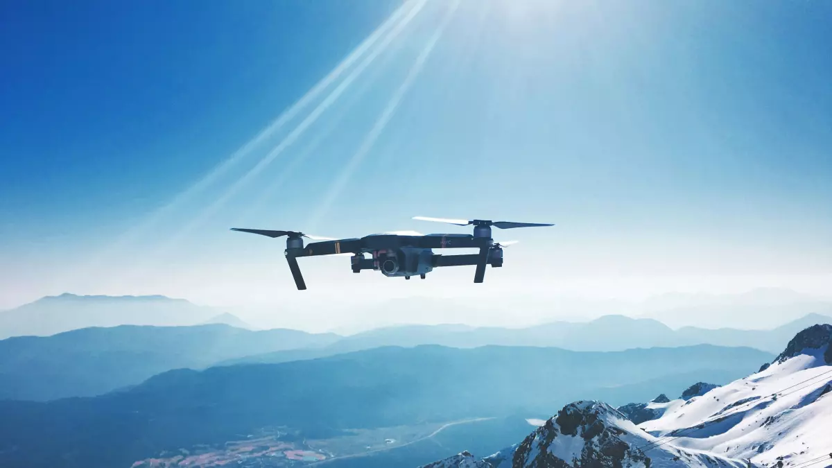 A drone flies high above a snowy mountain range, with a clear blue sky and sunbeams in the background.