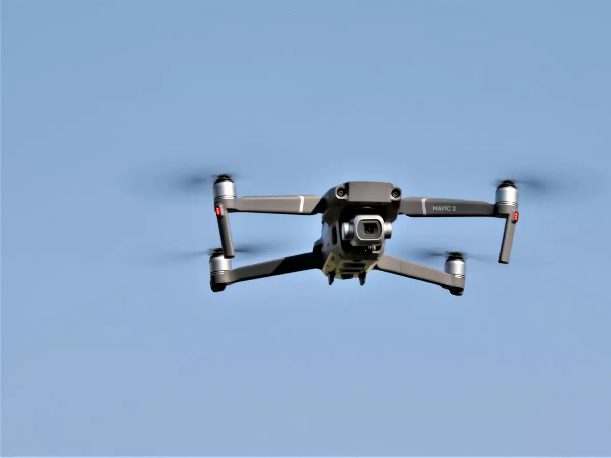 A drone is flying in the sky with a blue background. You can see the drone's sensors, propellers and its design.