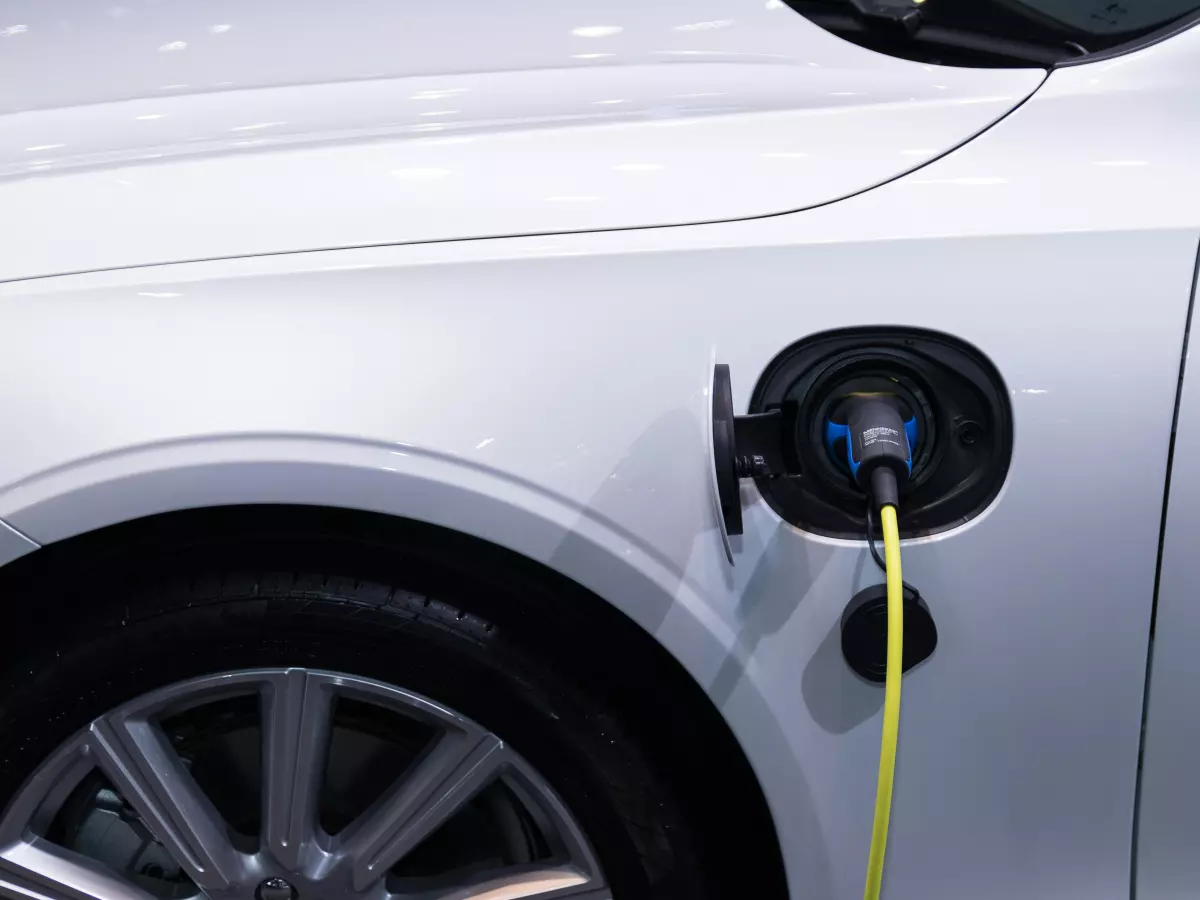 A close-up of a white electric vehicle being charged. The charging cable is plugged into the car's charging port, with the cable extending out of the frame.