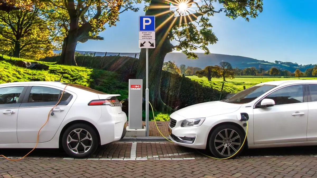 Two white electric cars plugged in to charge, one is a Chevrolet Volt and the other a Volvo, parked on a paved surface with a sign in the background.