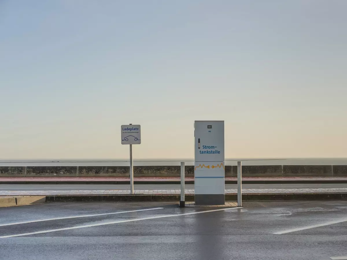 A charging station for electric vehicles is shown in the middle of a parking lot.  The station is a white box with a black sign that says "Strom-tankstelle." There are two signs on the left and right of the station that say "Ladeplatz."