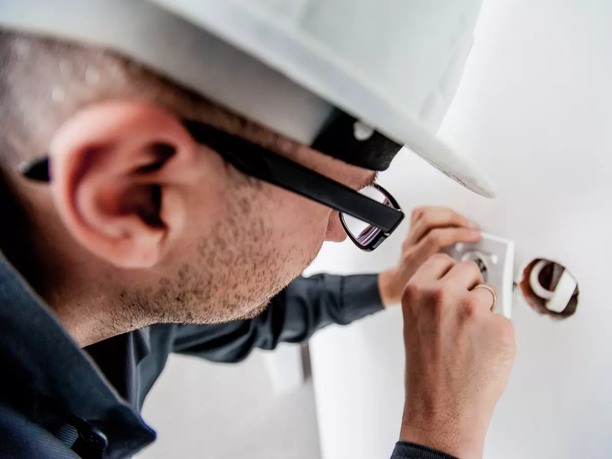 A man in a hard hat and glasses is working on an electrical outlet on a wall.