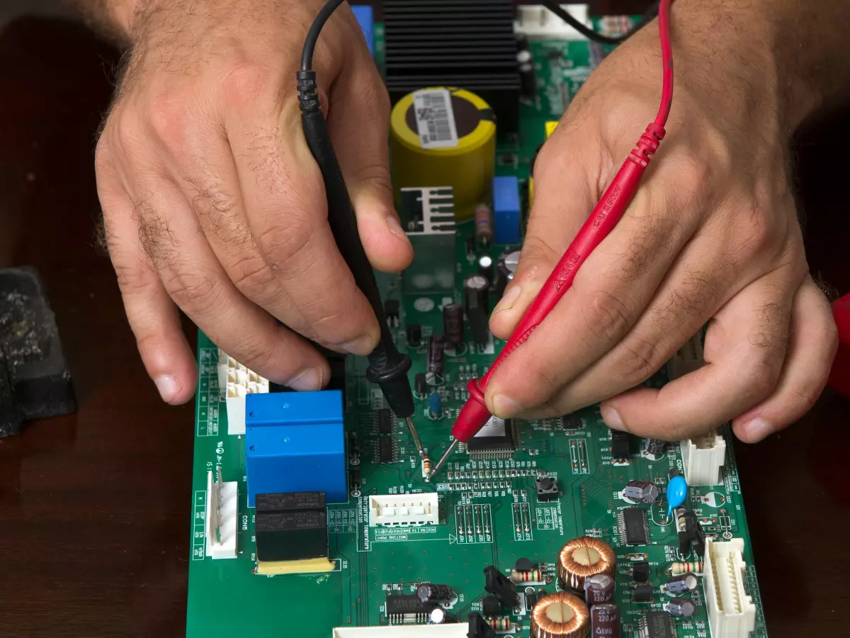 A technician is working on a printed circuit board (PCB). They are using a multimeter to test the board. The image is focused on the PCB and the technician's hands, with the background blurred.