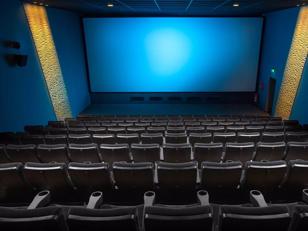 A wide shot of an empty movie theater, showcasing the large screen, rows of seats, and a blue wall behind the screen with golden stripes.