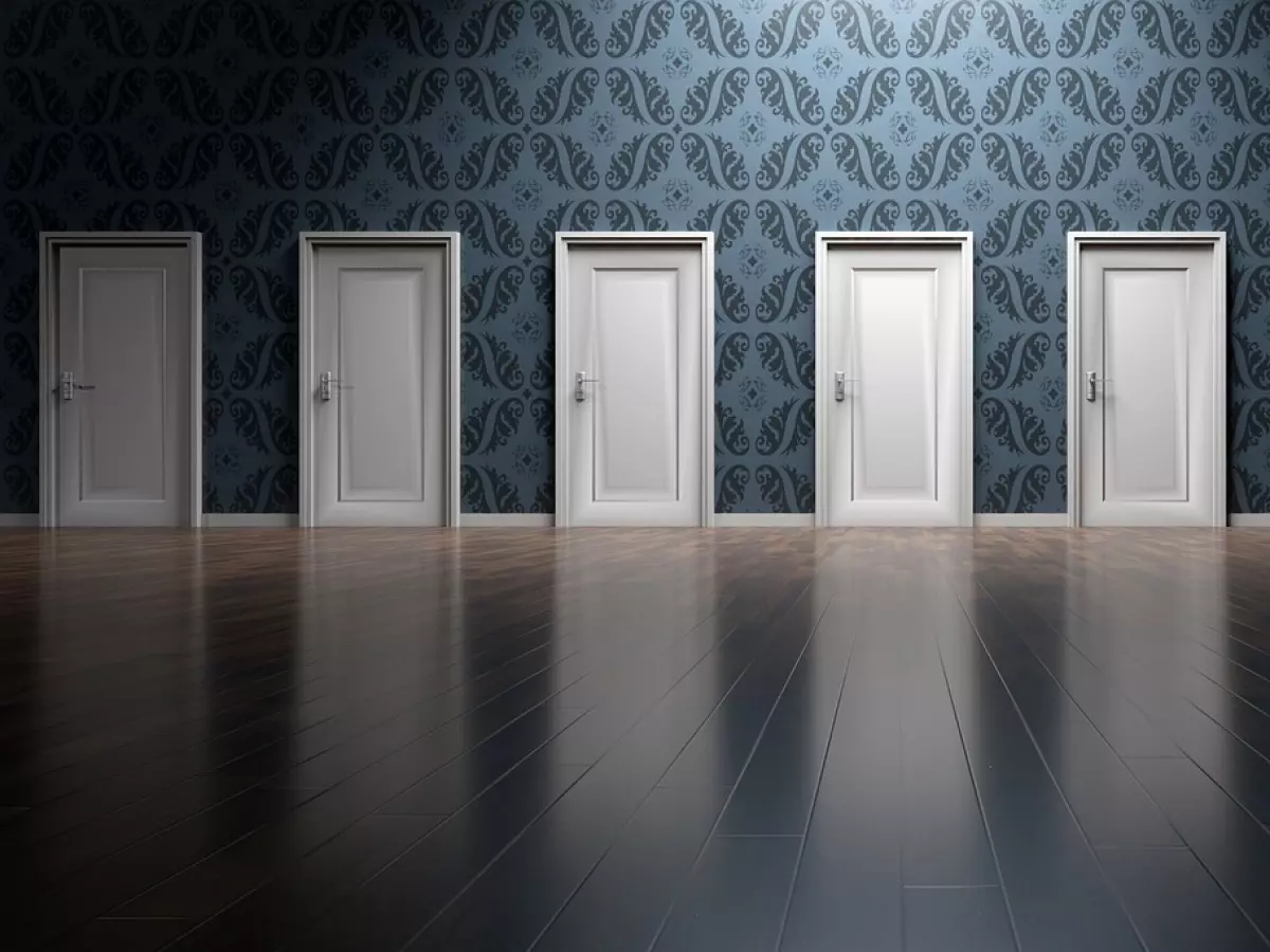 A row of seven closed white doors with brass handles, against a patterned wallpaper background. The doors are evenly spaced and lead into an empty room with a wooden floor.