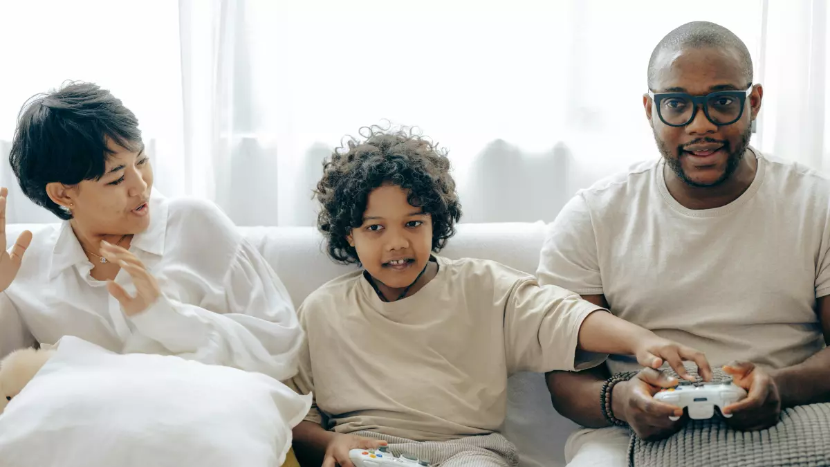 A father and his son are playing a video game together, with the father looking at the son with a smile.