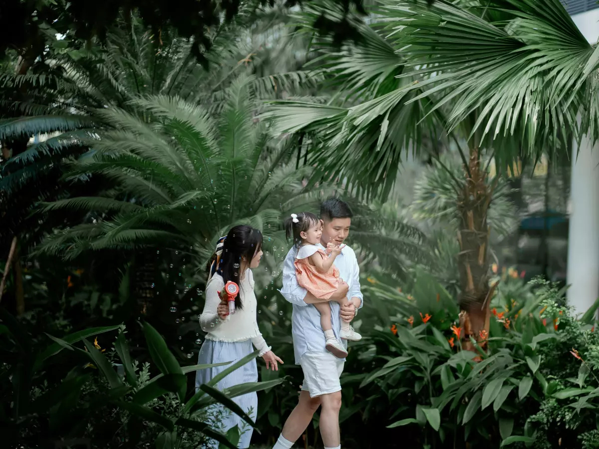 A young family is walking through a lush green garden. The father carries their daughter in his arms. The lush vegetation and warm colors create a sense of natural harmony.