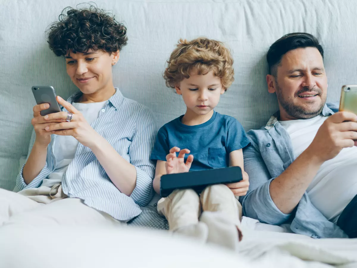 A family of three is sitting on a bed and looking at their phones. The mother is holding a phone in her hand, the father is holding a phone in his hand and the son is holding a tablet. They are all looking at their devices.