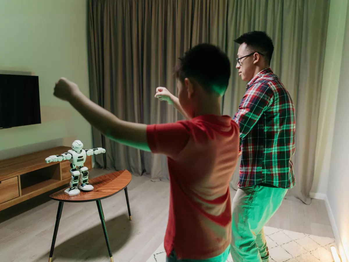 A father and son in a living room with a humanoid robot, the son is holding a smartphone, the robot is in the background