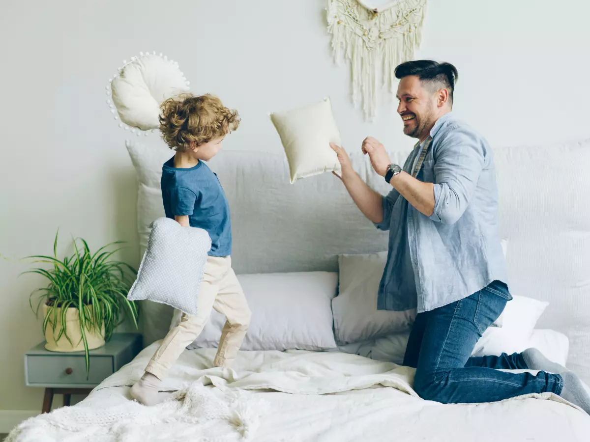 A father and son are playfully fighting on a bed, using pillows. The son is jumping in the air and holding a pillow in each hand, while the father is kneeling on the bed and holding a pillow in his right hand. The father's expression is one of amusement and the son's expression is one of playful aggression.