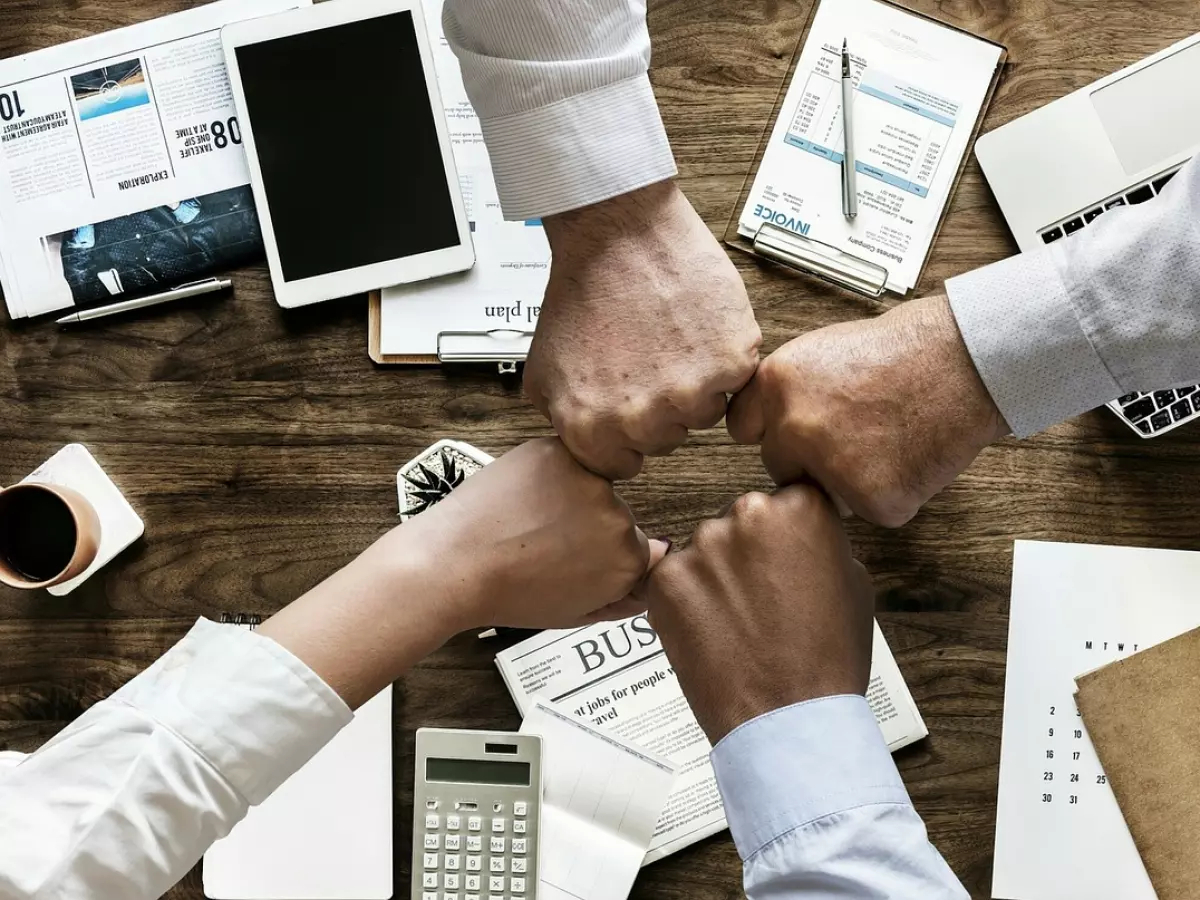 Four hands clasped together in the center of a desk, surrounded by office supplies, symbolizing teamwork and collaboration.