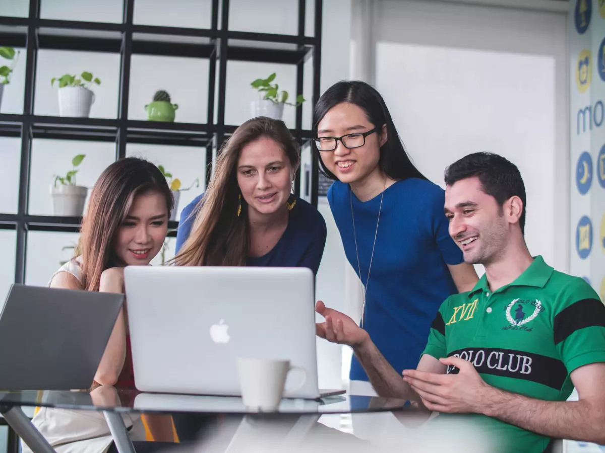 Four people are looking at a laptop. They are sitting at a table. The people are all smiling. There are plants in the background.