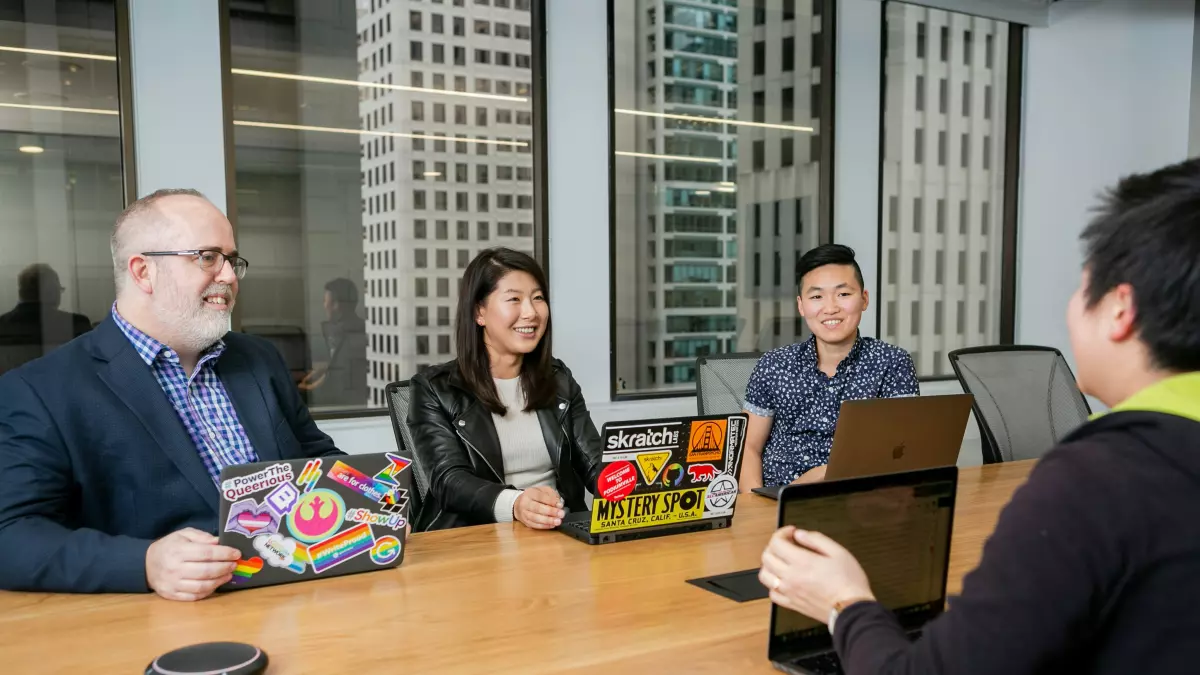 Four people are sitting around a table in a meeting room, discussing something on their laptops.