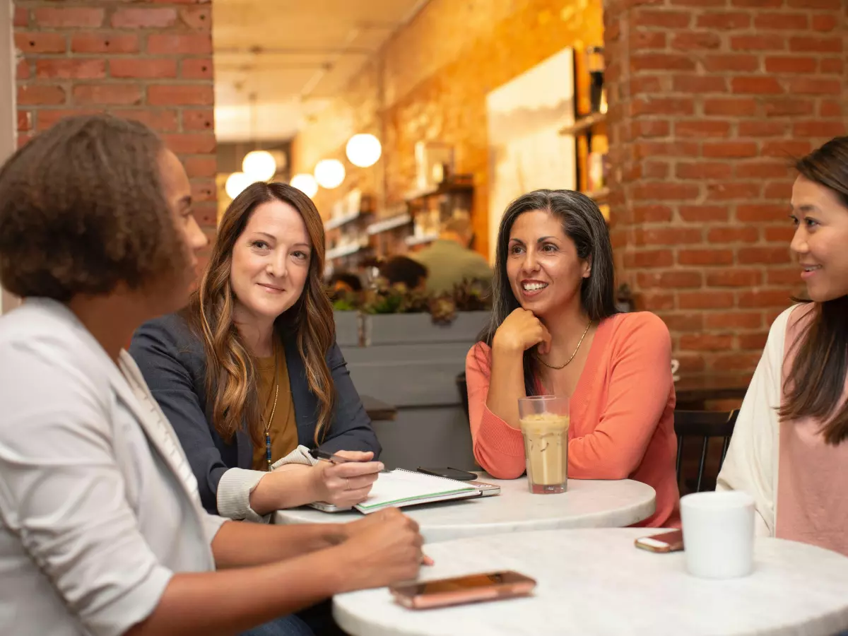 Four people, three women and one man, are sitting at a table in a cafe, talking and looking at each other. The cafe is decorated with exposed brick walls and warm lighting. The people appear to be casually dressed, and they have a relaxed and friendly atmosphere. The image is well-lit, and the composition is balanced.