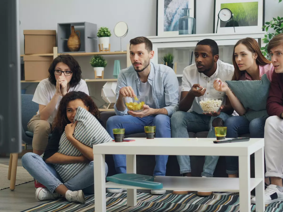 A group of friends watching TV together. They are all excited and engaged in what they are watching. There are snacks and drinks on the table in front of them. 