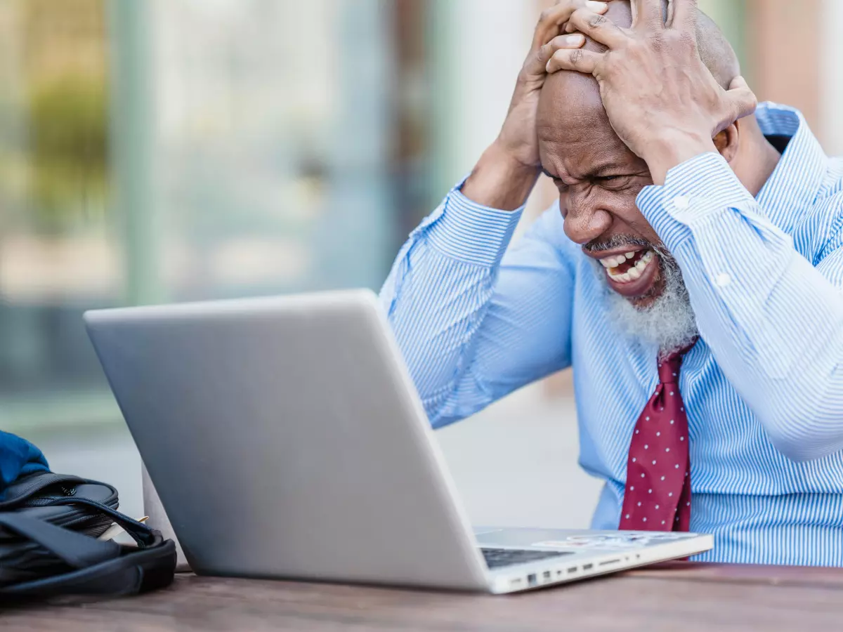 A man in a suit and tie sitting at a table, looking distressed as he looks at a laptop screen.