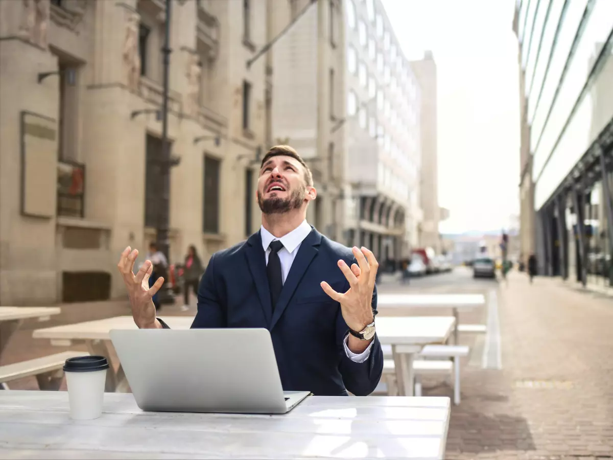 A man in a suit sits at a table outside, looking distressed with his hands raised in the air. He is looking up at the sky in exasperation. He has a laptop in front of him.