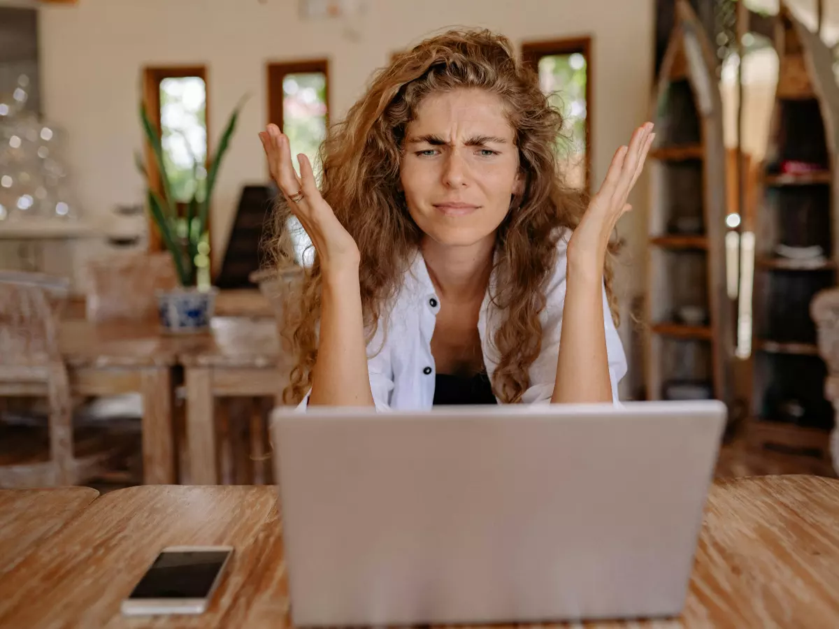 A woman sits at a wooden table, looking frustrated as she works on a laptop. Her arms are raised in a gesture of exasperation. A smartphone lies on the table beside the laptop.